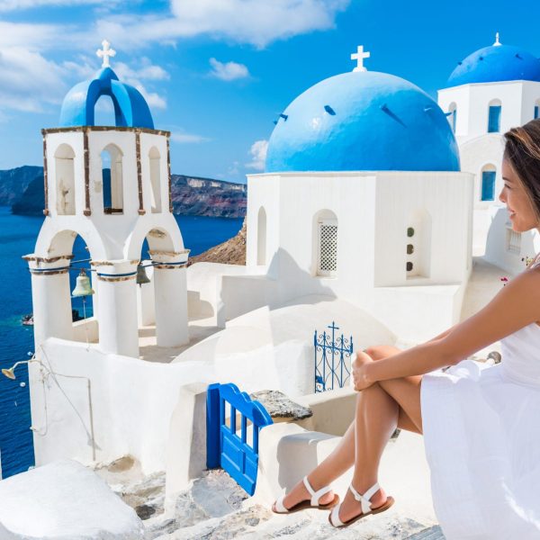 Luxury travel Greece vacation Asian elegant woman on honeymoon enjoying view of blue sea and three domes in white dress. Summer holidays on Oia, Santorini greek island, Europe.