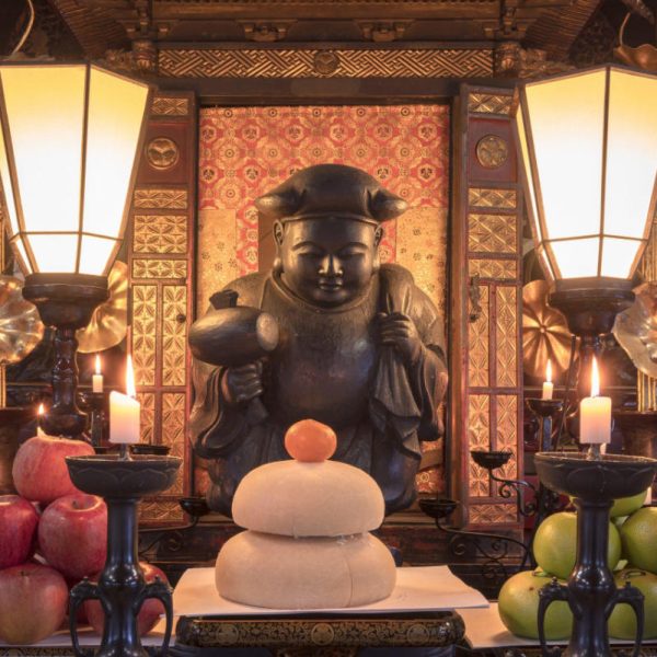 tokyo, japan - january 07 2022: New year's mochi rice, clementine and apples offerings in front of a wooden statue depicting one of the Seven Lucky Gods, Daikokuten, the god of wealth, household and kitchen holding a golden mallet of fortune seated on ba